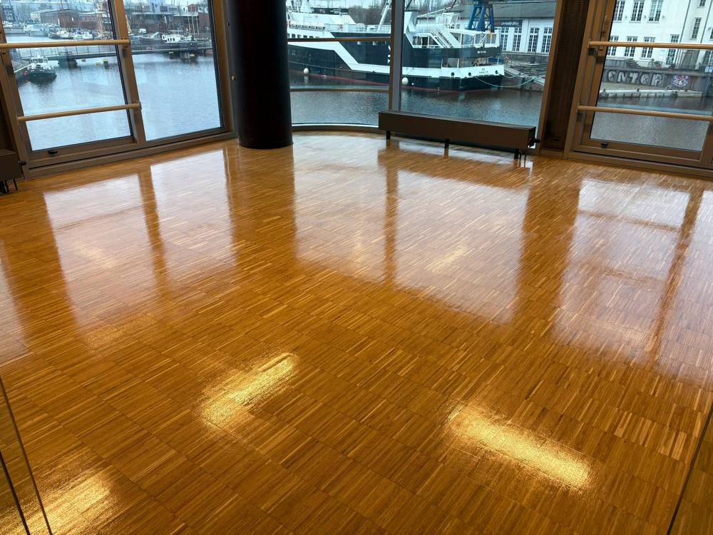 Ganz neu verlegtes, glänzendes Parkett in hellem Raum vor großen Panoramafenstern mit Hafenblick