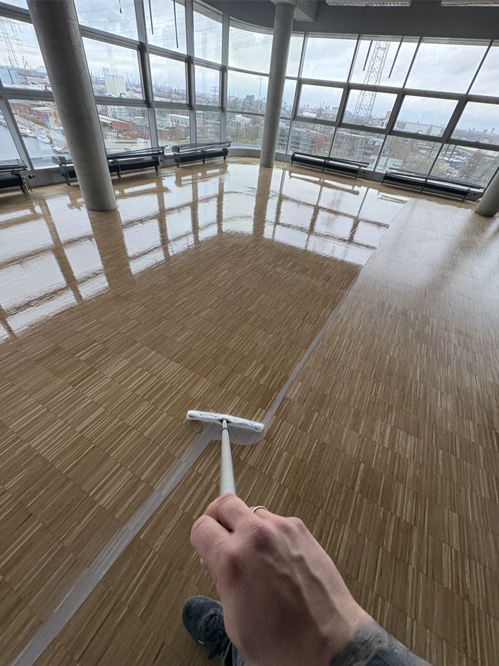 Auftragen von Bodenpflege auf Holzparkett in hellem Geschäftsraum mit Panorama-Fenstern