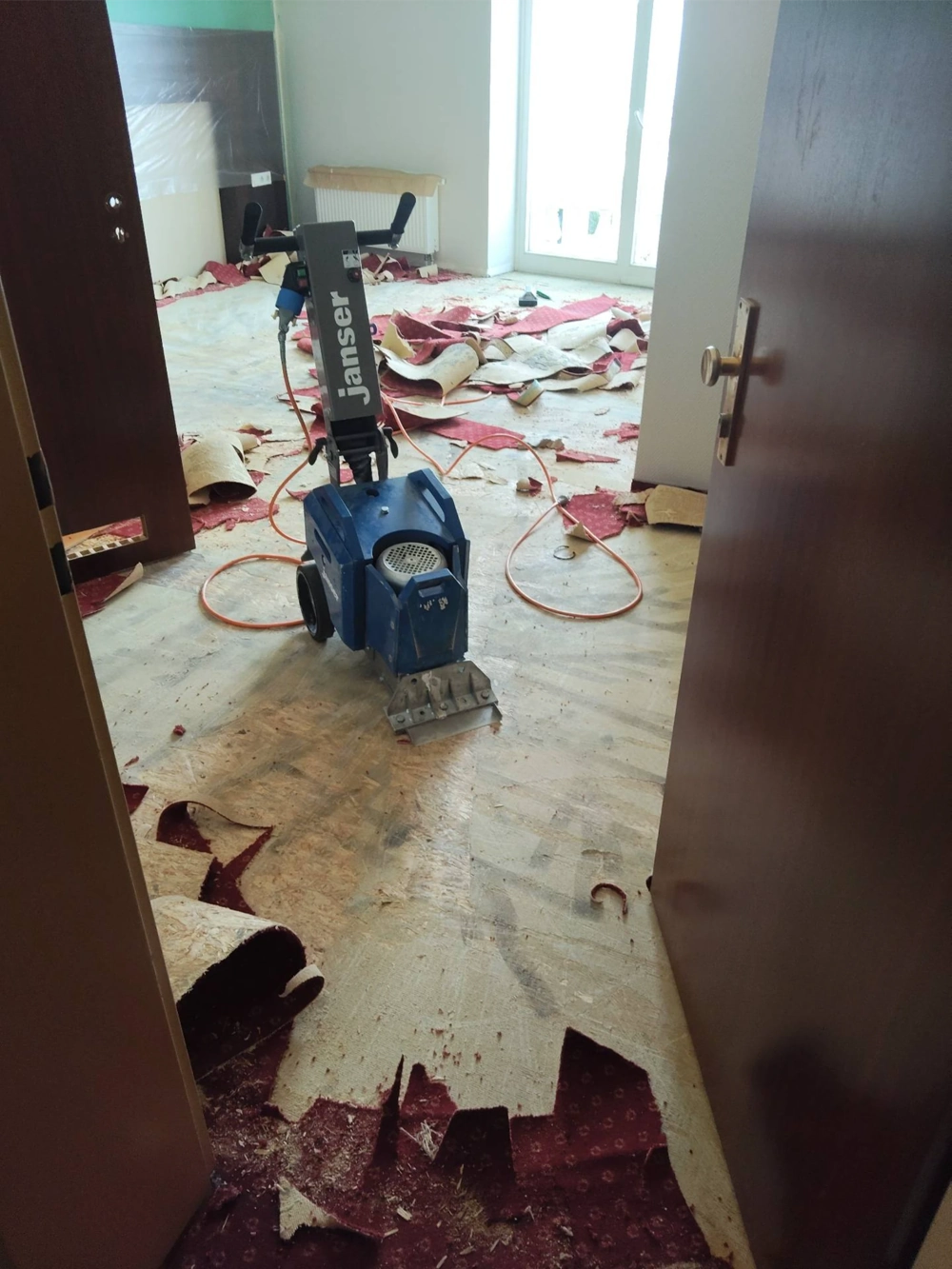 Maschinelle Teppichentfernung: aufgerauter Boden mit Kabeln und roten Teppichresten im Raum