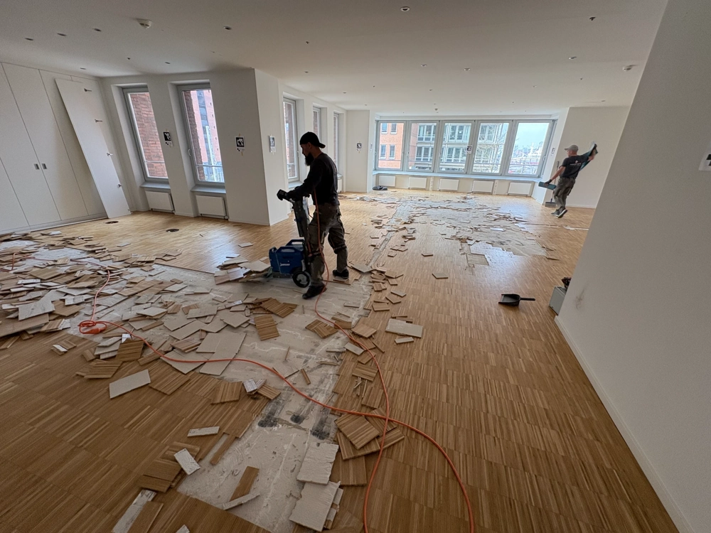 Zwei Handwerker beim Parkett entfernen in hellem Großraumbüro, Holzstäbe auf dem Boden verteilt und Bodenentfernungsgerät im Einsatz