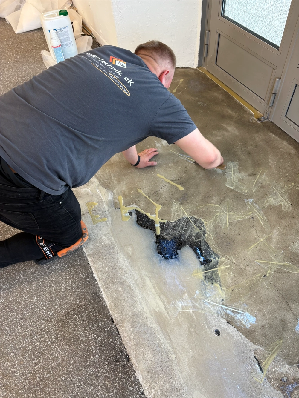Handwerker kniet auf rohem Betonboden und entfernt alte Klebereste – Untergrund vorbereiten