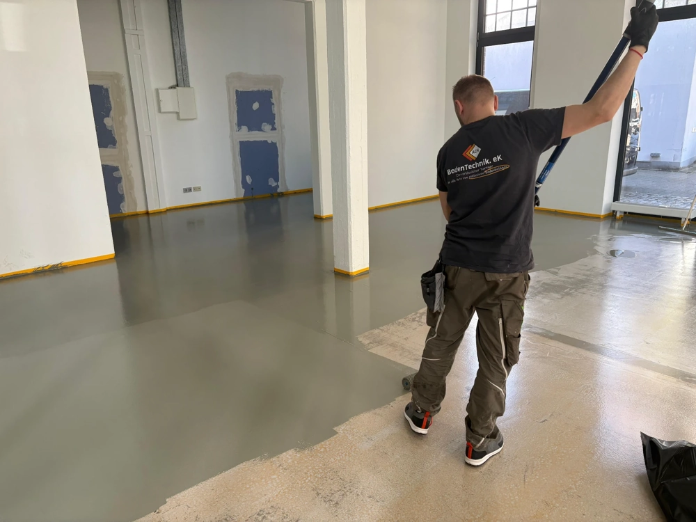 Handwerker trägt Epoxidharzboden in großem gewerblichen Raum mit langem Rakel auf ebenen Betonuntergrund auf
